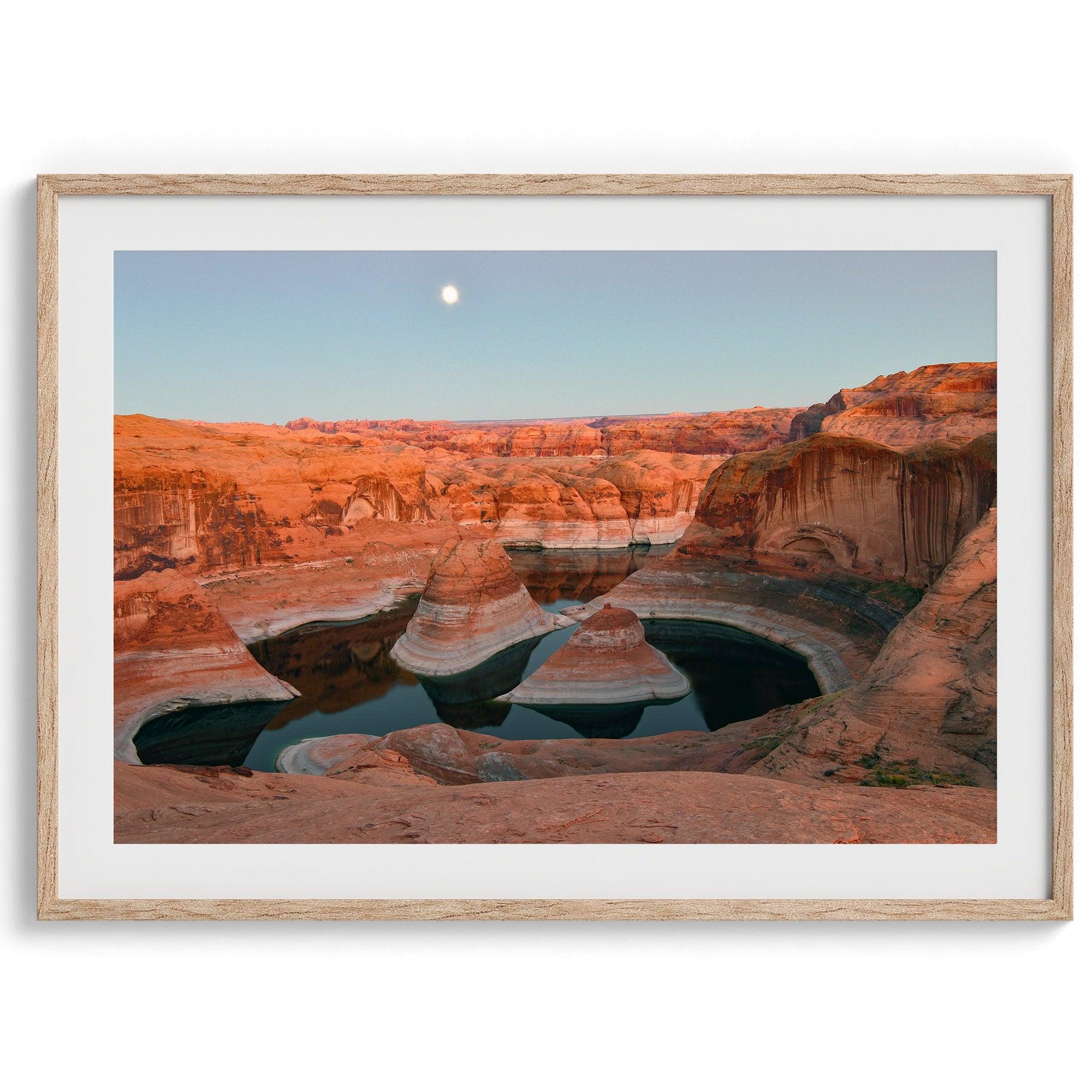 Landscape photo of Reflection Canyon in the Utah desert at sunset, with fiery red hues reflected in still water below a blue sky with a moon.