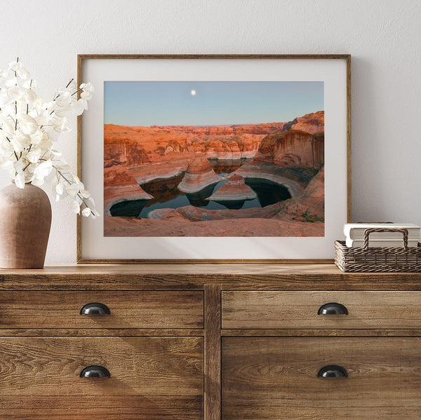 Landscape photo of Reflection Canyon in the Utah desert at sunset, with fiery red hues reflected in still water below a blue sky with a moon.