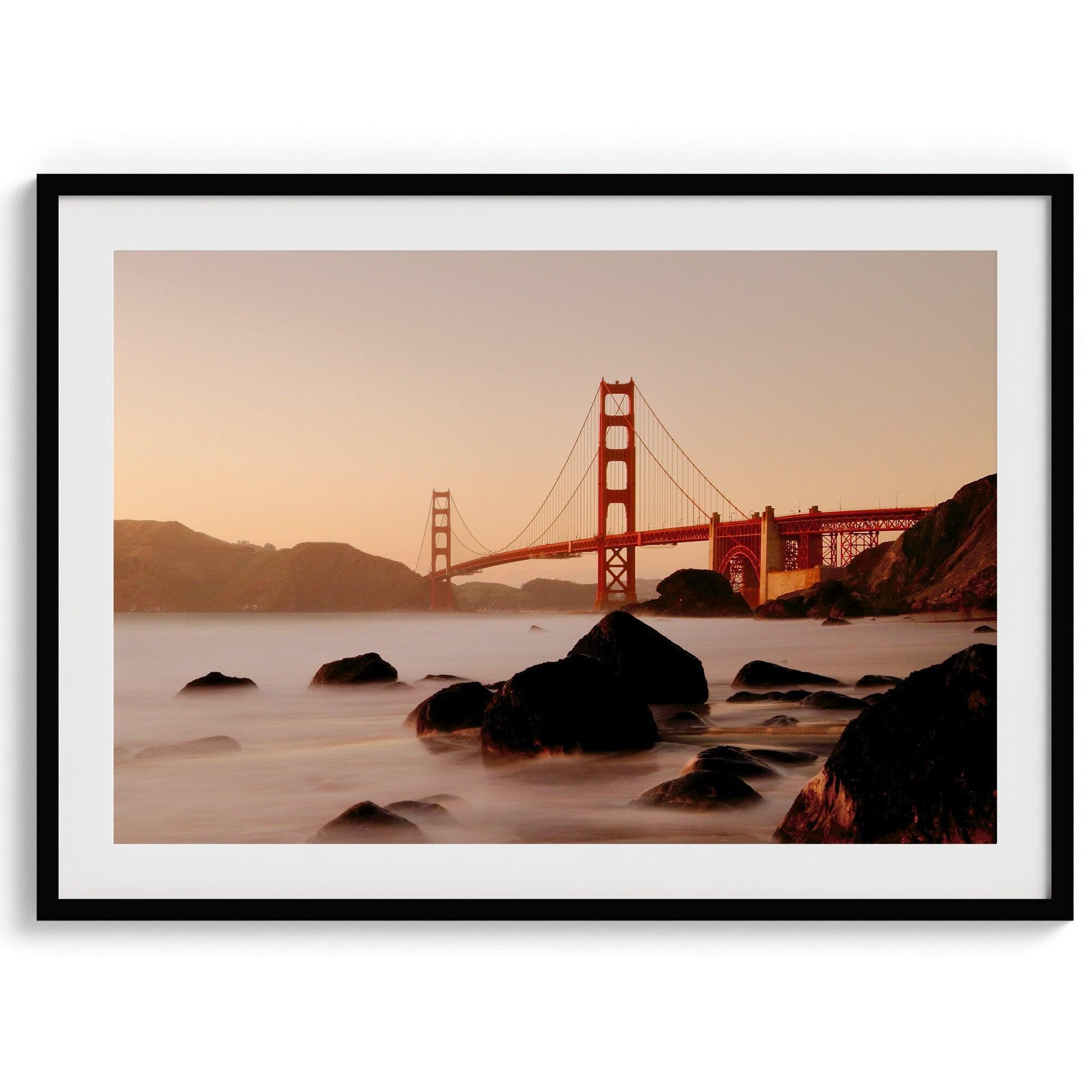 Limited-edition print: Golden Gate Bridge at sunset. Long exposure blurs ocean waves, contrasting with fiery red & orange sky. Dramatic scene with bridge framed by foreground rocks.