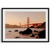 Limited-edition print: Golden Gate Bridge at sunset. Long exposure blurs ocean waves, contrasting with fiery red & orange sky. Dramatic scene with bridge framed by foreground rocks.