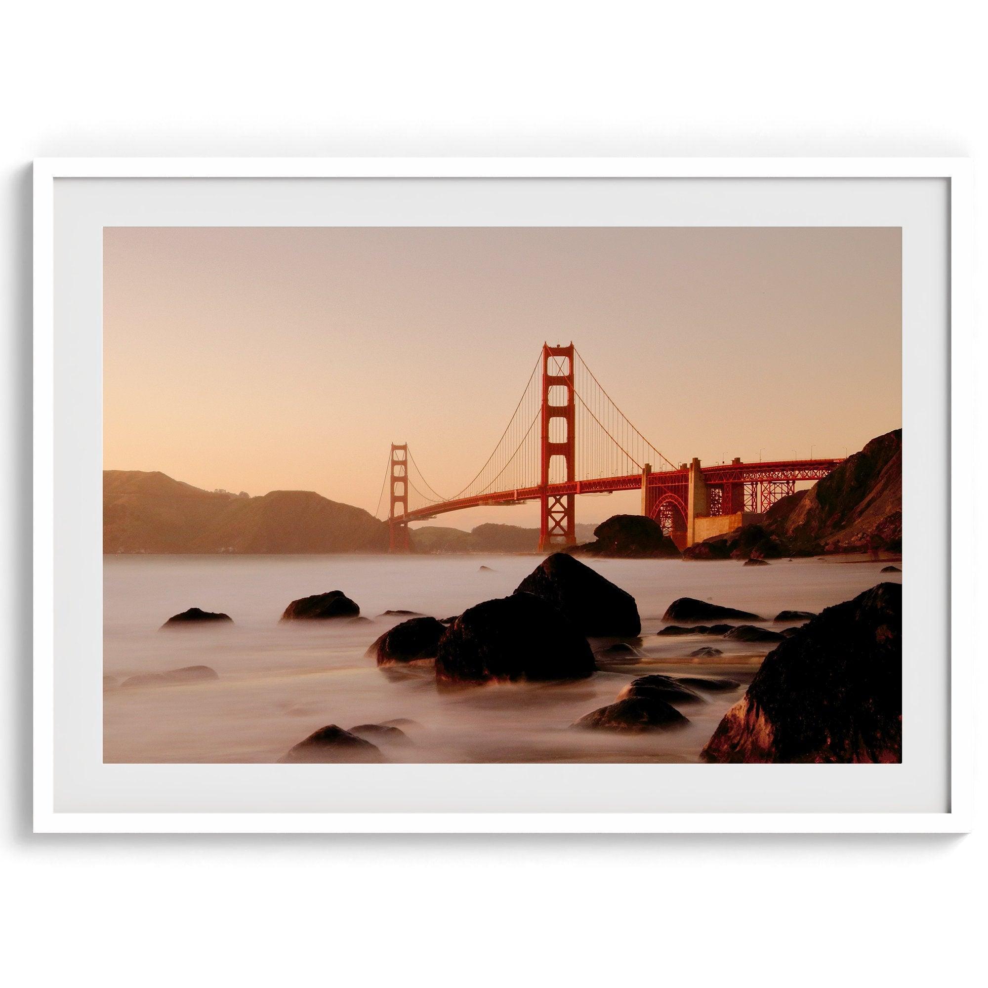 Limited-edition print: Golden Gate Bridge at sunset. Long exposure blurs ocean waves, contrasting with fiery red & orange sky. Dramatic scene with bridge framed by foreground rocks.