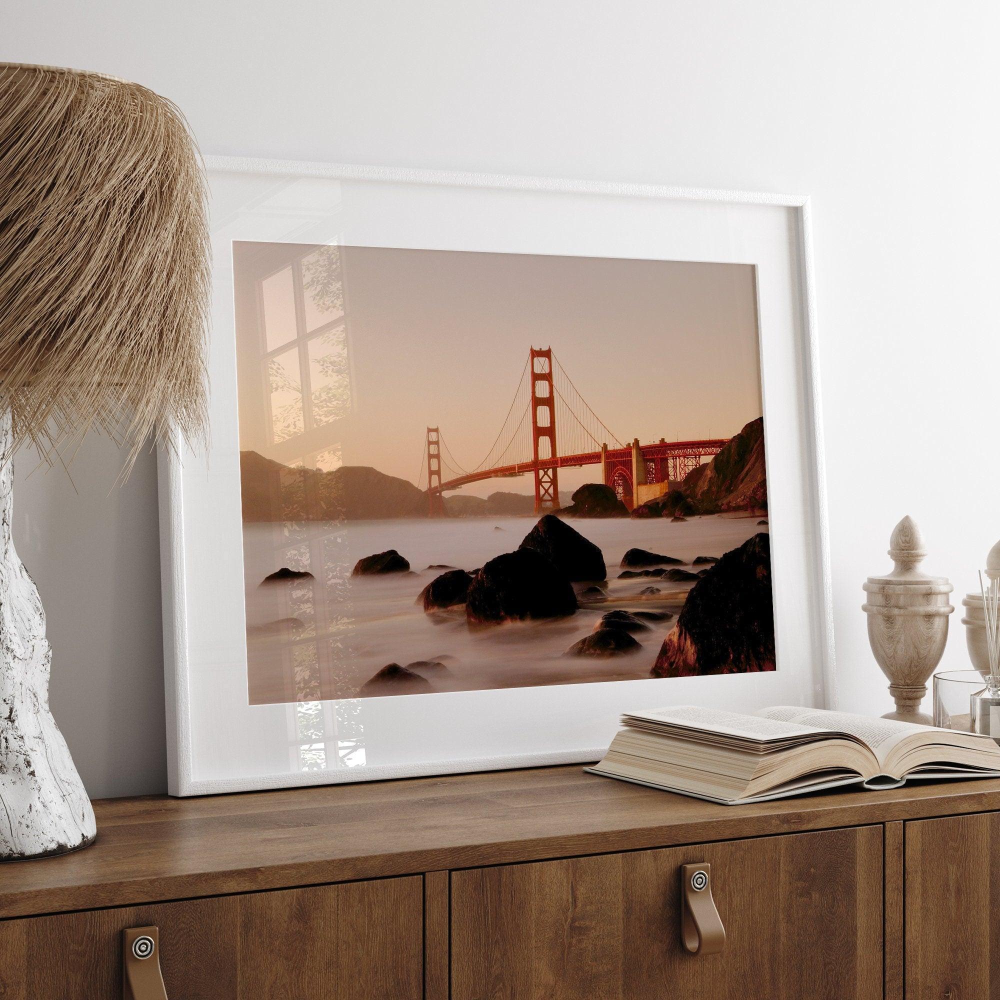 Limited-edition print: Golden Gate Bridge at sunset. Long exposure blurs ocean waves, contrasting with fiery red & orange sky. Dramatic scene with bridge framed by foreground rocks.