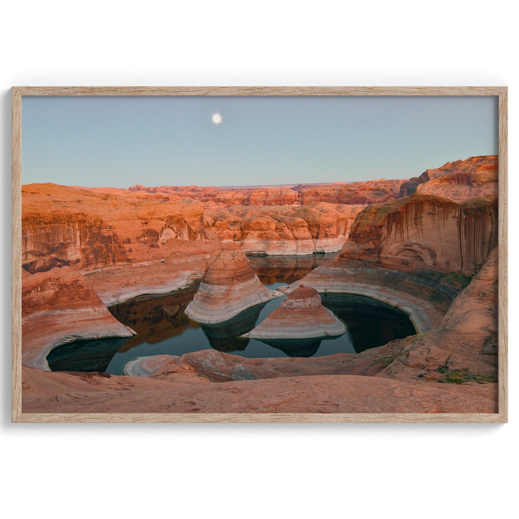 Landscape photo of Reflection Canyon in the Utah desert at sunset, with fiery red hues reflected in still water below a blue sky with a moon.