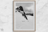 A fine art print showing five skiers traversing a vast, snowy mountain landscape under a pale winter sky, surrounded by untouched wilderness.