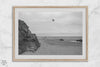 Black-and-white framed photo of a kite flying over a quiet California beach with rocky cliffs and an overcast sky.