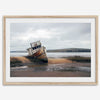 Weathered shipwreck resting on a quiet coastal shore, surrounded by marshland and a moody, overcast sky.