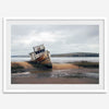 Weathered shipwreck resting on a quiet coastal shore, surrounded by marshland and a moody, overcast sky.