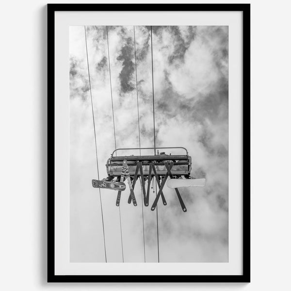 Monochrome ski lift view from below with hanging skis and cloudy mountain sky background.