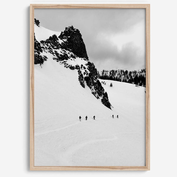 A fine art print showing five skiers traversing a vast, snowy mountain landscape under a pale winter sky, surrounded by untouched wilderness.