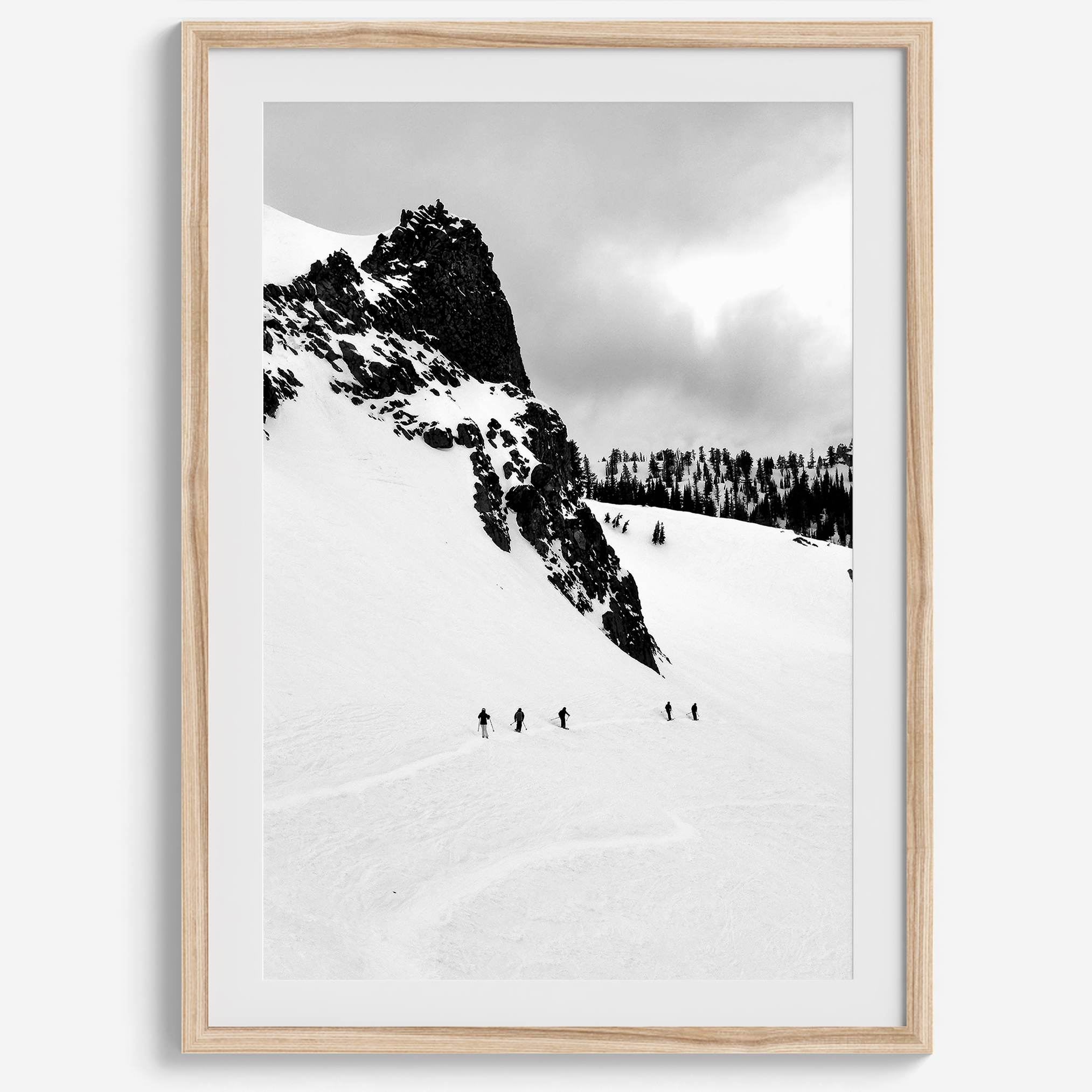 A fine art print showing five skiers traversing a vast, snowy mountain landscape under a pale winter sky, surrounded by untouched wilderness.
