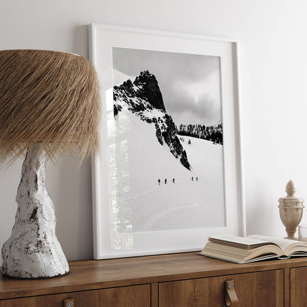 A fine art print showing five skiers traversing a vast, snowy mountain landscape under a pale winter sky, surrounded by untouched wilderness.