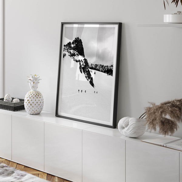A fine art print showing five skiers traversing a vast, snowy mountain landscape under a pale winter sky, surrounded by untouched wilderness.