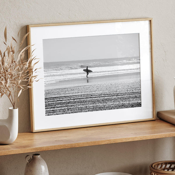 Black and white surfer photography showing a lone surfer on the beach, with waves in the background, capturing minimalist coastal beauty.