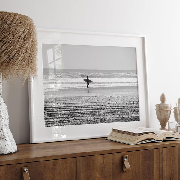 Black and white surfer photography showing a lone surfer on the beach, with waves in the background, capturing minimalist coastal beauty.