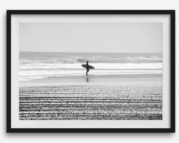 Black and white surfer photography showing a lone surfer on the beach, with waves in the background, capturing minimalist coastal beauty.