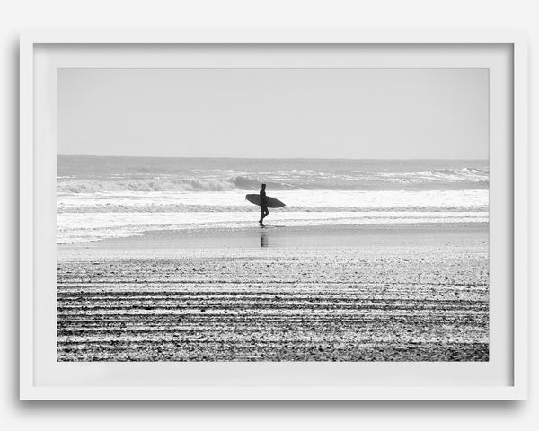 Black and white surfer photography showing a lone surfer on the beach, with waves in the background, capturing minimalist coastal beauty.
