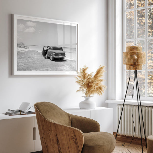 Black and white vintage beach print featuring a classic car parked at the edge of the ocean, capturing a retro coastal scene with timeless charm.