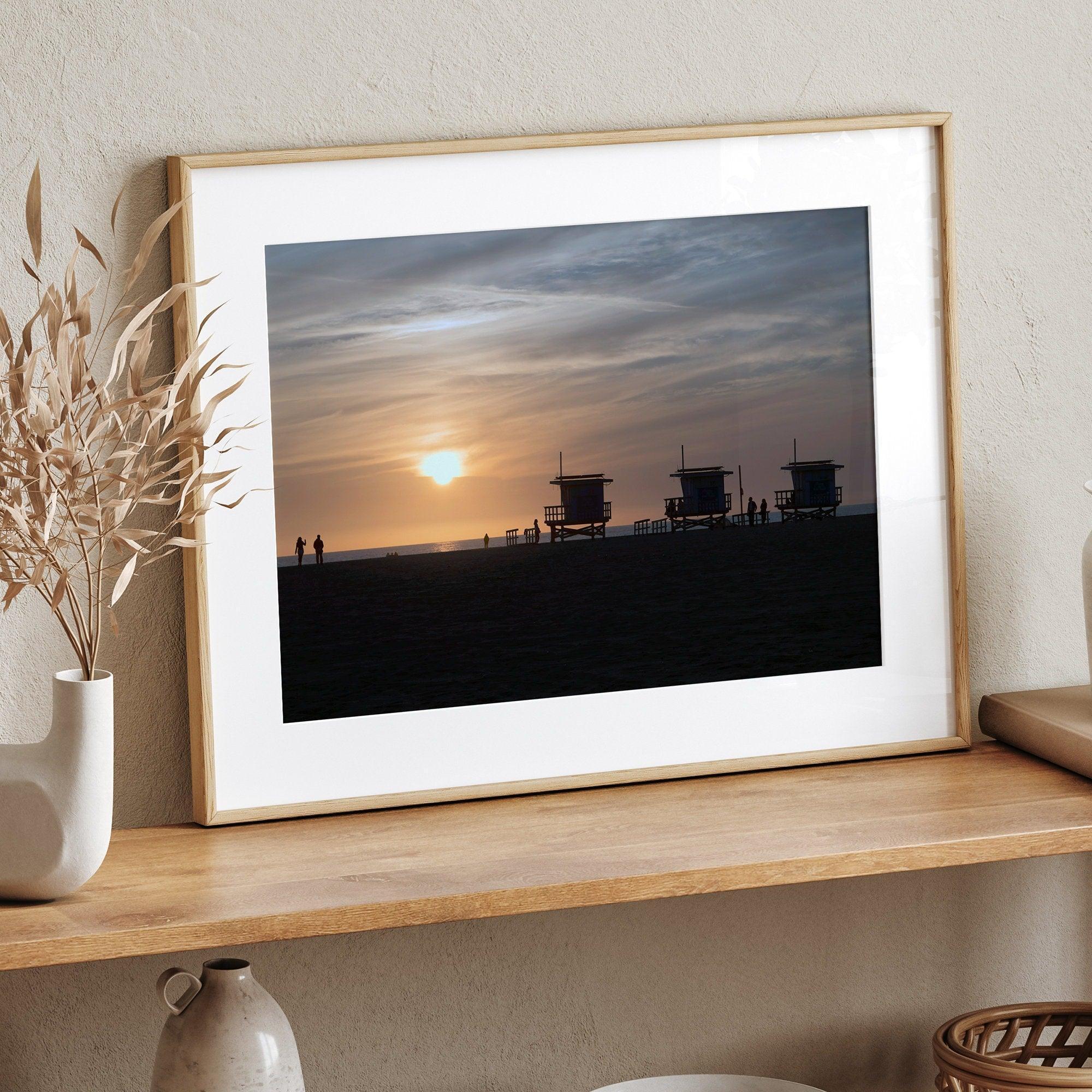 Step into Venice Beach with our fine art beach sunset framed or unframed print. The print showcases the beach in sunset with the silhouettes of the famous lifeguard towers and people in the sunset.