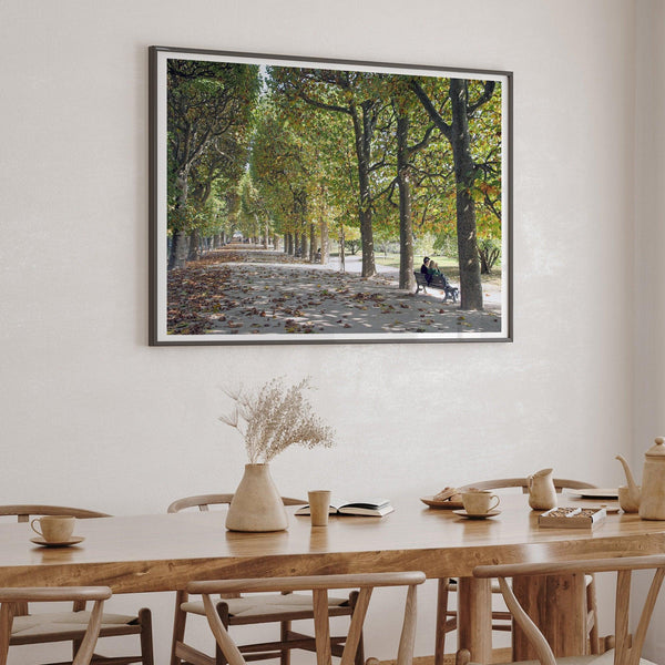 A fine art Paris print of a Parisian garden in fall showcasing a couple sitting on a bench under the fall color trees with foliage all around.