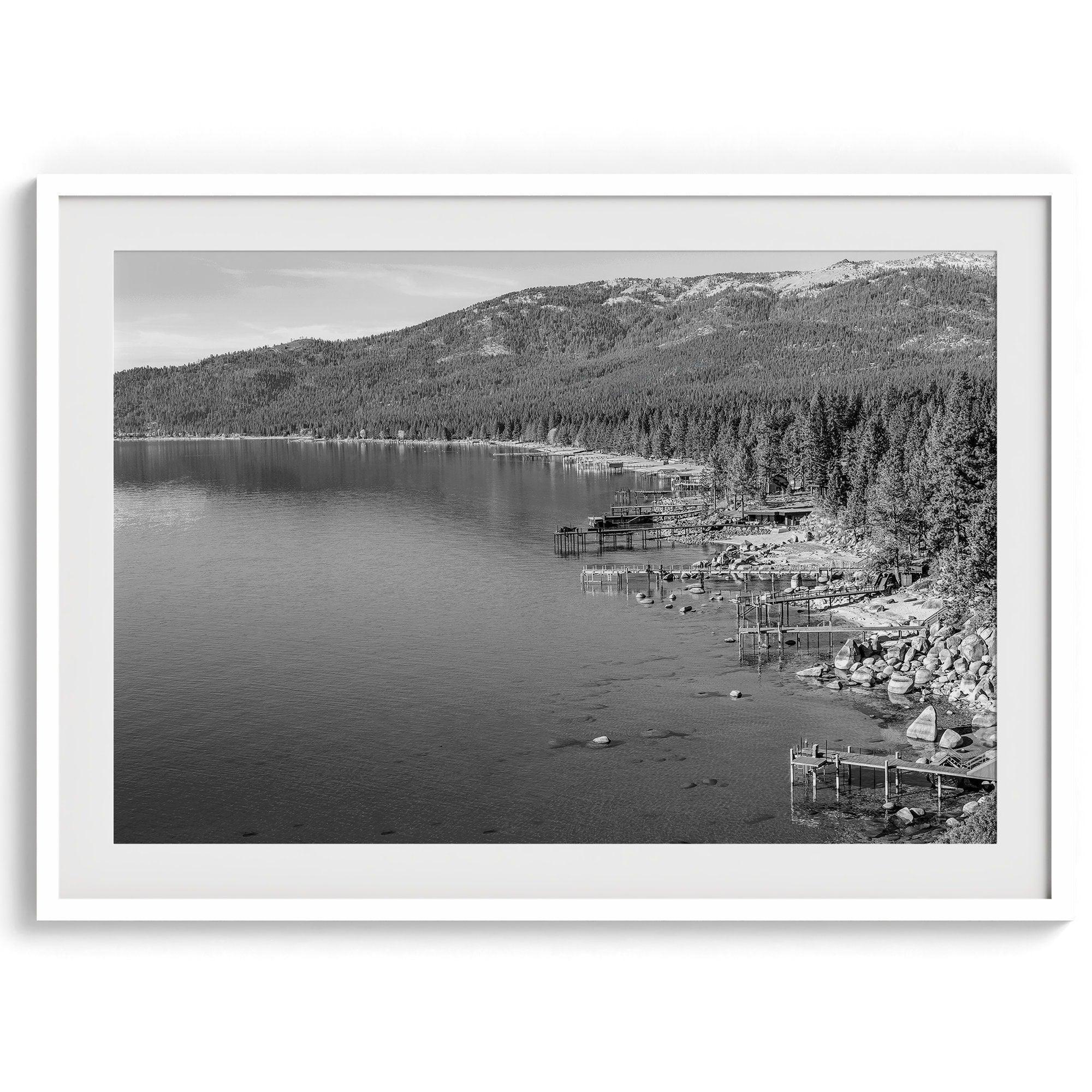 A fine art black and white Lake Tahoe print showcasing docks and boats nested on the lake waters on the left, while the beachfront, nestled against a backdrop of a lush forest and majestic mountain, paints a picturesque scene on the right.