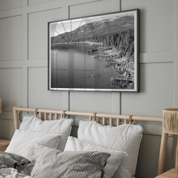 A fine art black and white Lake Tahoe print showcasing docks and boats nested on the lake waters on the left, while the beachfront, nestled against a backdrop of a lush forest and majestic mountain, paints a picturesque scene on the right.