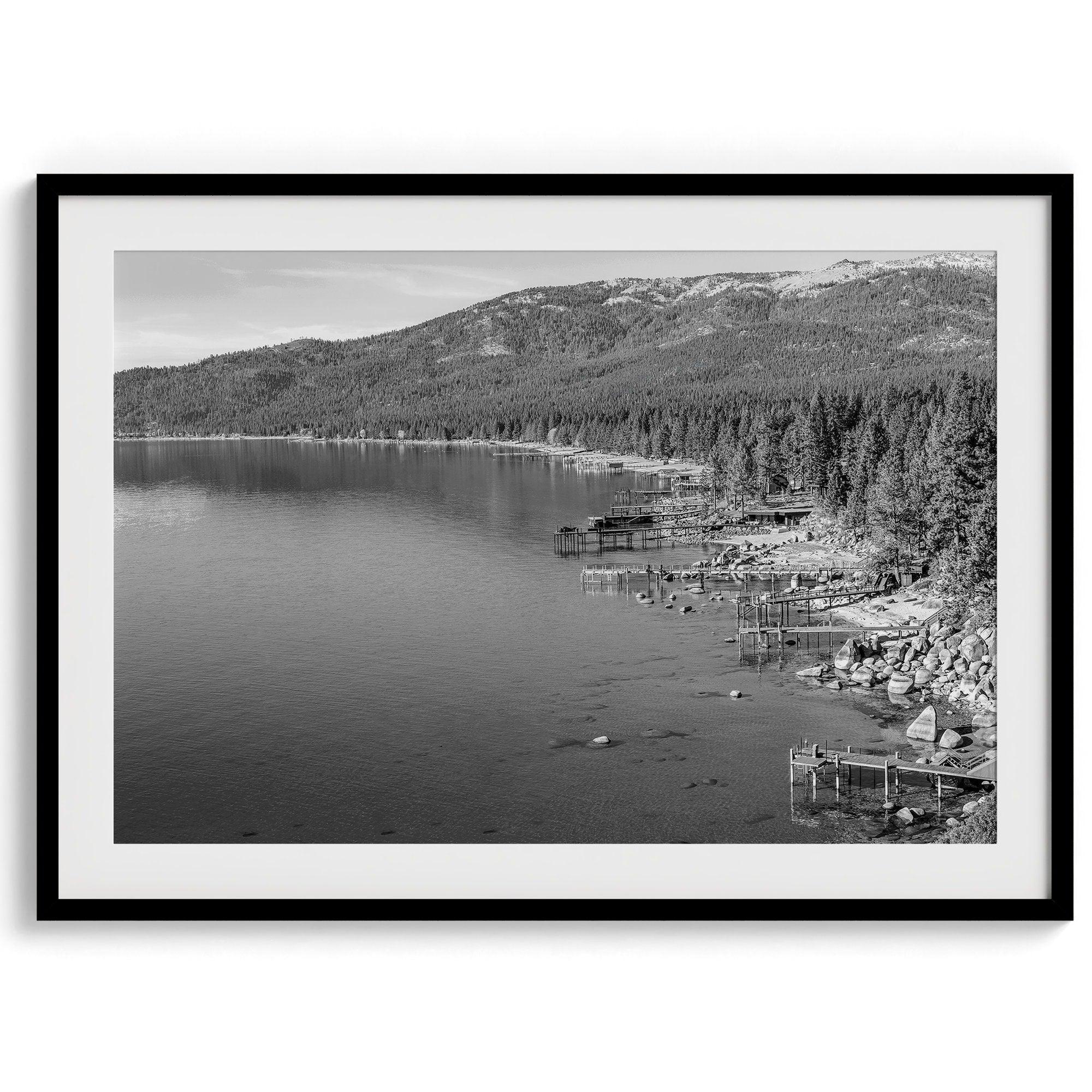 A fine art black and white Lake Tahoe print showcasing docks and boats nested on the lake waters on the left, while the beachfront, nestled against a backdrop of a lush forest and majestic mountain, paints a picturesque scene on the right.