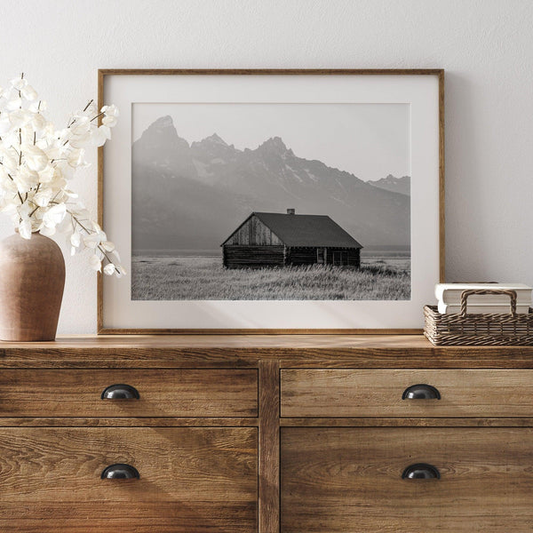 Step into Grand Teton National Park with this fine art black and white mountain print. This western landscape photo showcases a century-old rustic hut at the foot of the snowy, pointy Teton mountains.