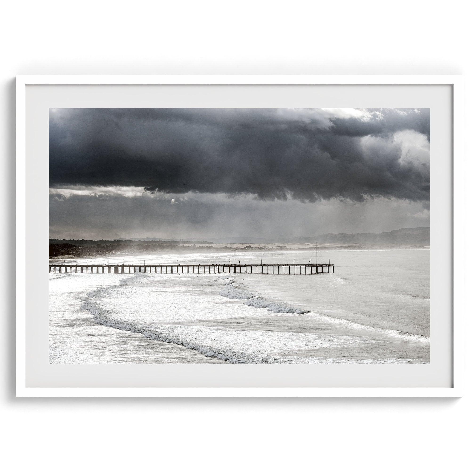 This fine art ocean photography print was taken in Pismo Beach, California, and showcases a distant view of an ocean pier and stormy clouds. Looking long and hard, you can see tens of surfers around the pier.