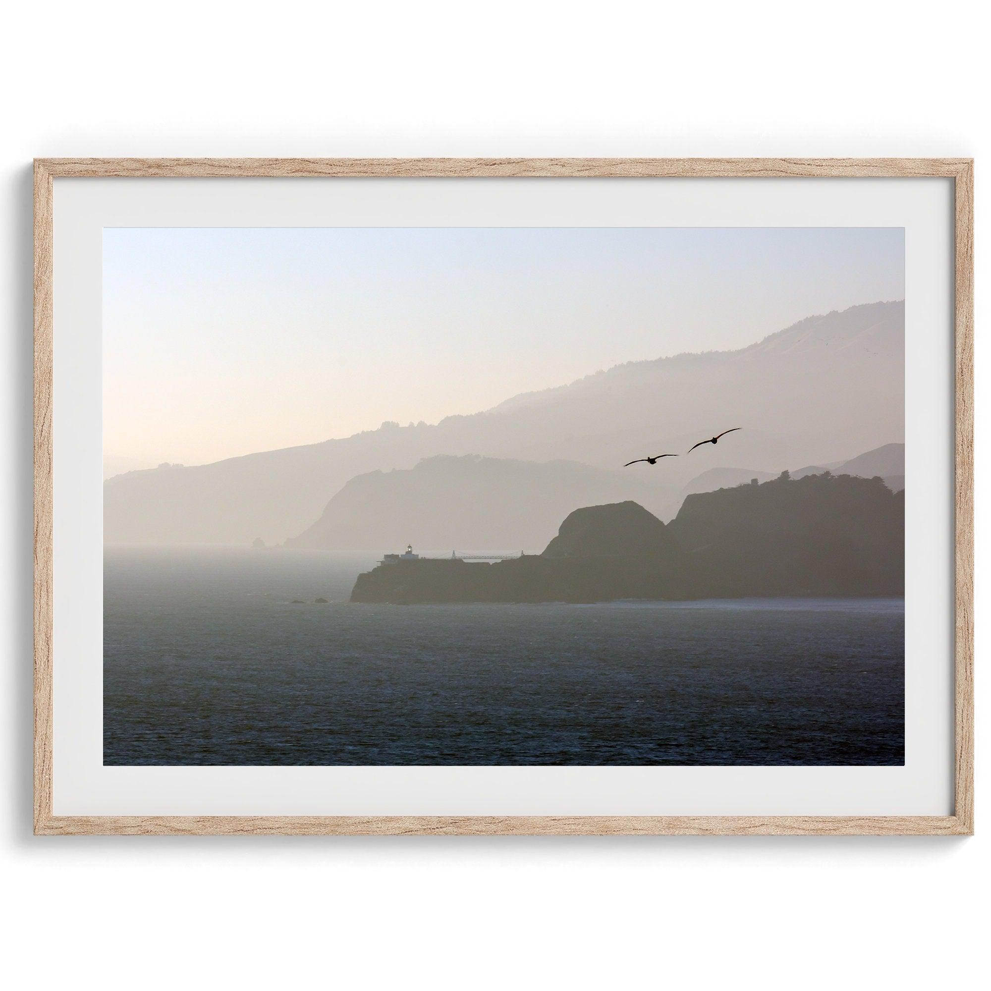 This fine art coastal photography print showcases two birds majestically flying over the Pacific Ocean towards the layered Marin mountains in the sunset. On one of the ridges, there is a lighthouse. The photo was taken from Lands End San Francisco.