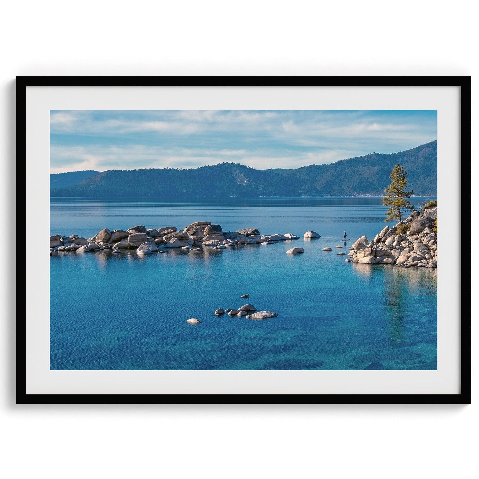 A fine art Lake Tahoe print showcasing a lone stand-up paddle peacefully gliding across the clear water lake near the huge rocks of Sand Harbor Beach with the backdrop of the Sierra Nevada mountains.