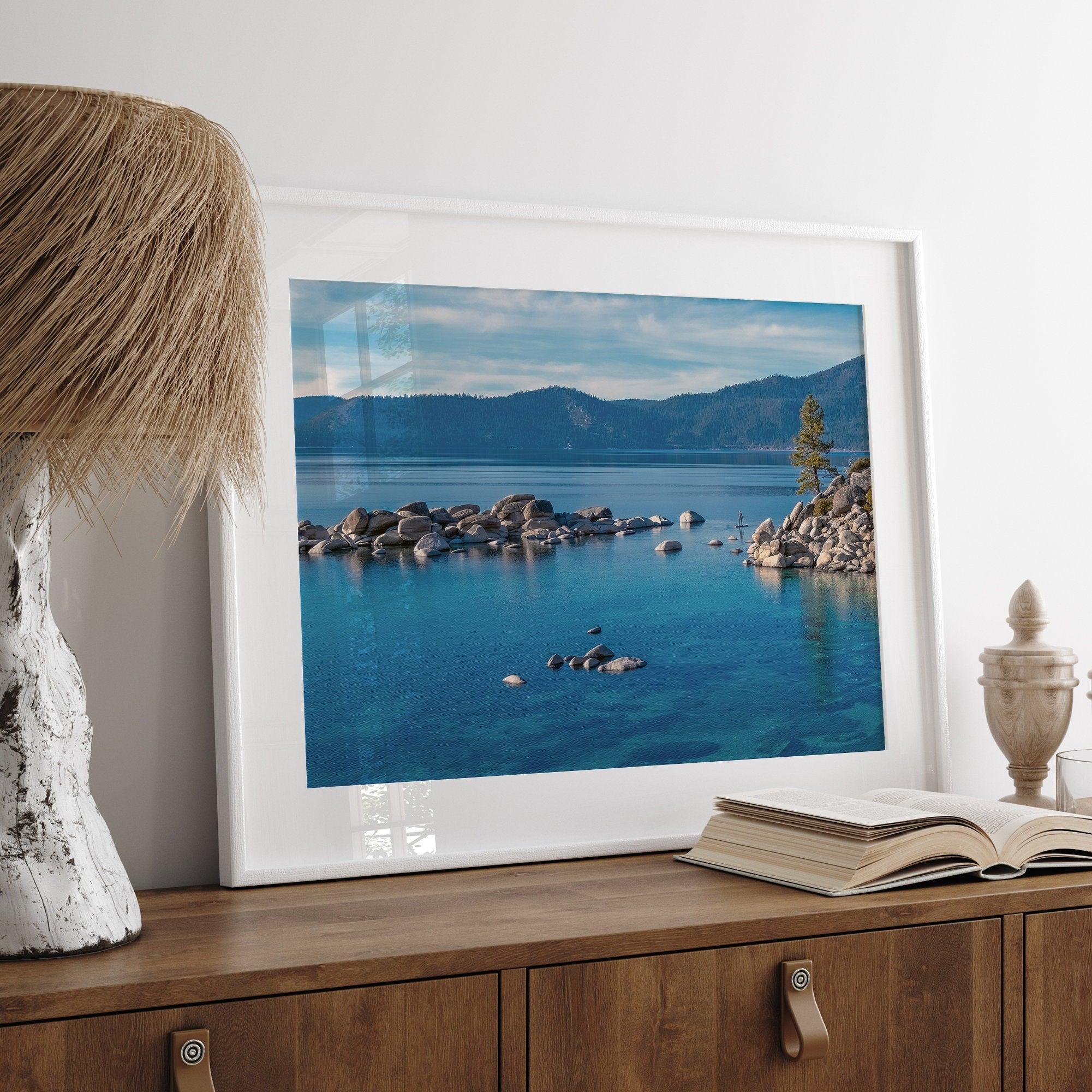 A fine art Lake Tahoe print showcasing a lone stand-up paddle peacefully gliding across the clear water lake near the huge rocks of Sand Harbor Beach with the backdrop of the Sierra Nevada mountains.