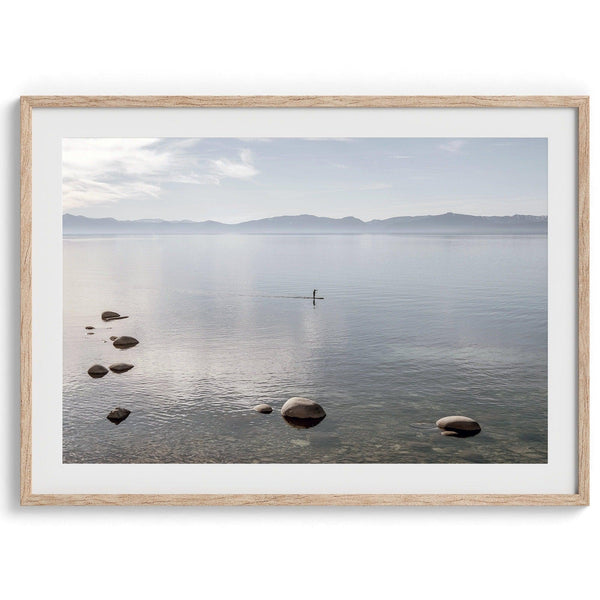 Fine art photography print of Lake Tahoe featuring a serene blue water surrounded by Pacific Northwest mountains, with a lone stand-up paddle silhouette on the lake.