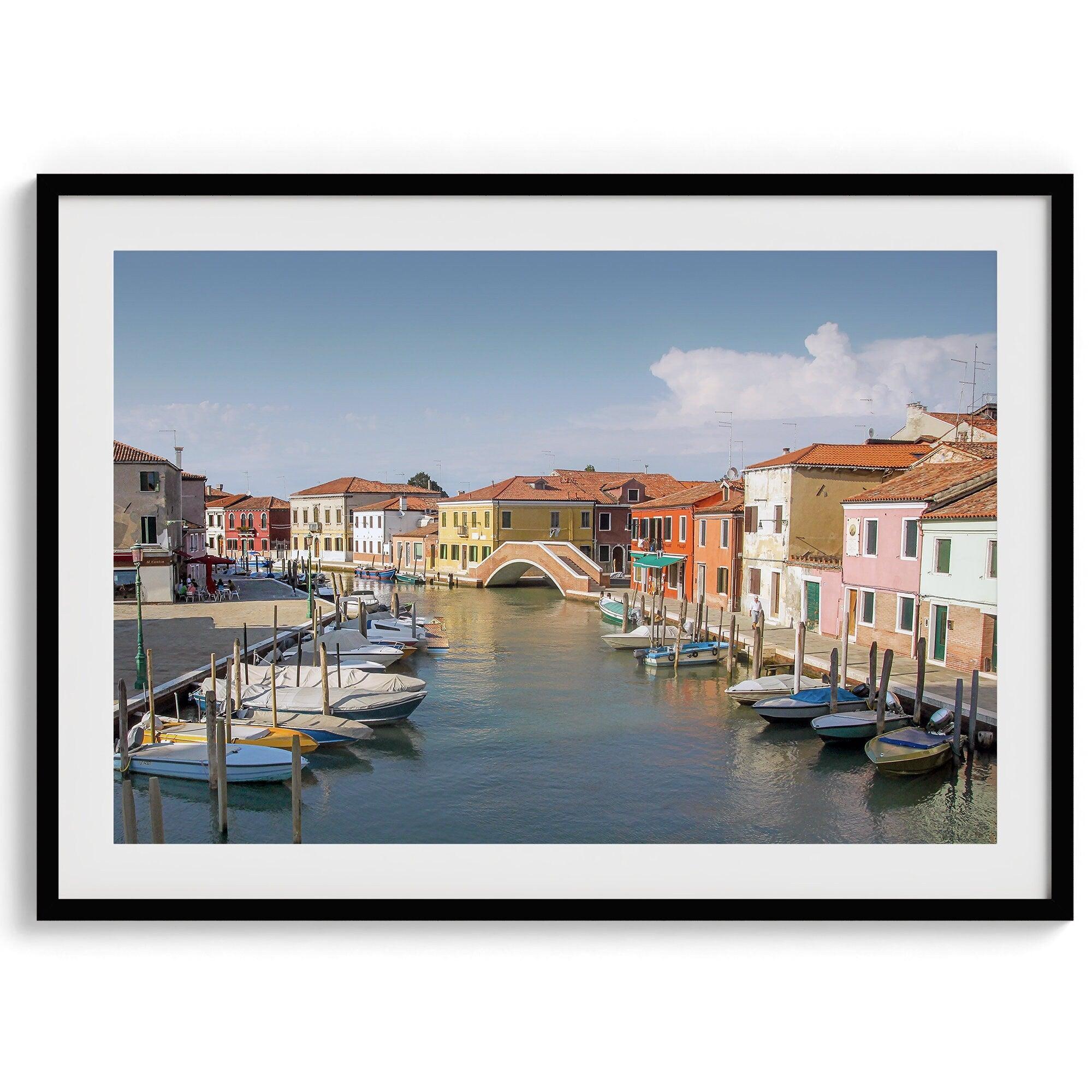 A stunning framed Venice print. The picturesque vibrant streets of Murano are lined with houses in an array of vibrant colors. The sun casts a beautiful glow on the tranquil canal, which adds to the charm of this peaceful Italian town.