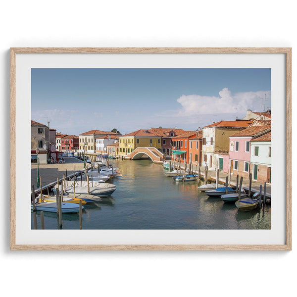 A stunning framed Venice print. The picturesque vibrant streets of Murano are lined with houses in an array of vibrant colors. The sun casts a beautiful glow on the tranquil canal, which adds to the charm of this peaceful Italian town.