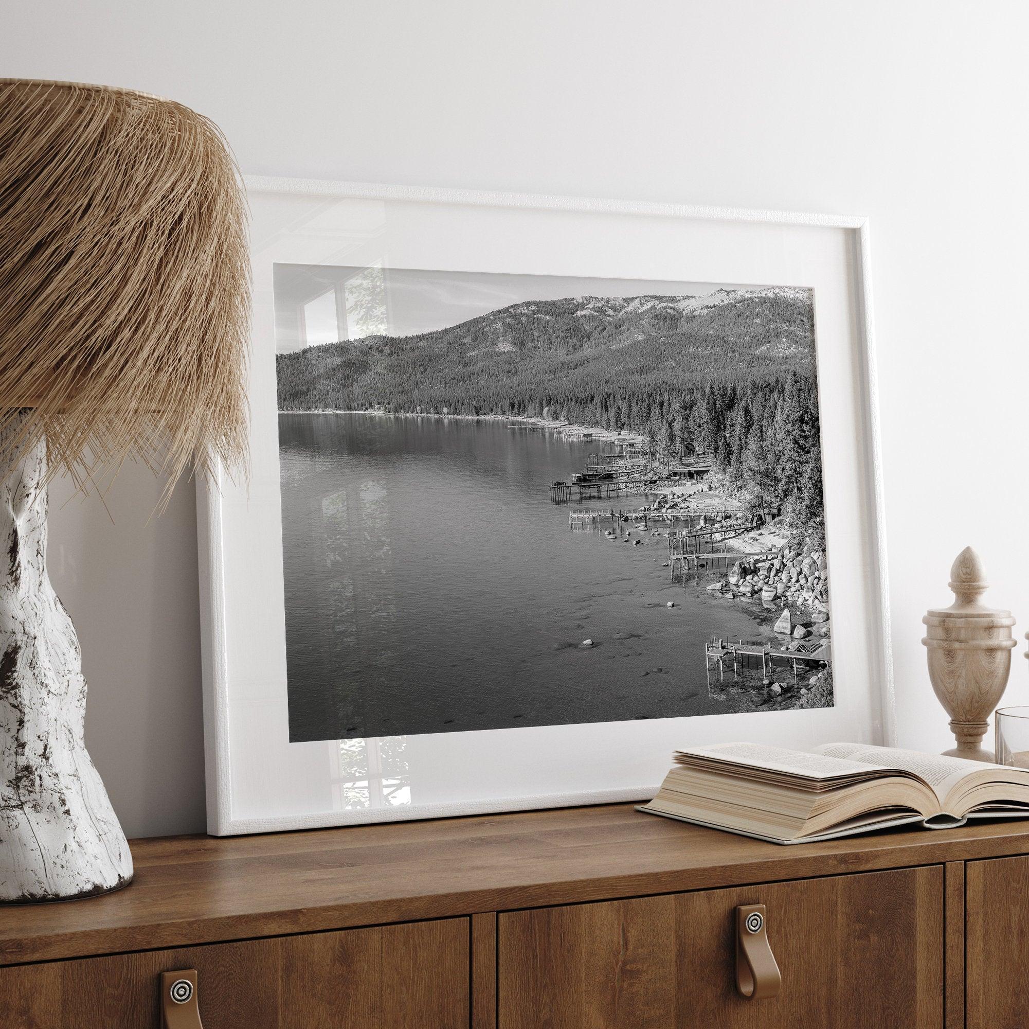 A fine art black and white Lake Tahoe print showcasing docks and boats nested on the lake waters on the left, while the beachfront, nestled against a backdrop of a lush forest and majestic mountain, paints a picturesque scene on the right.