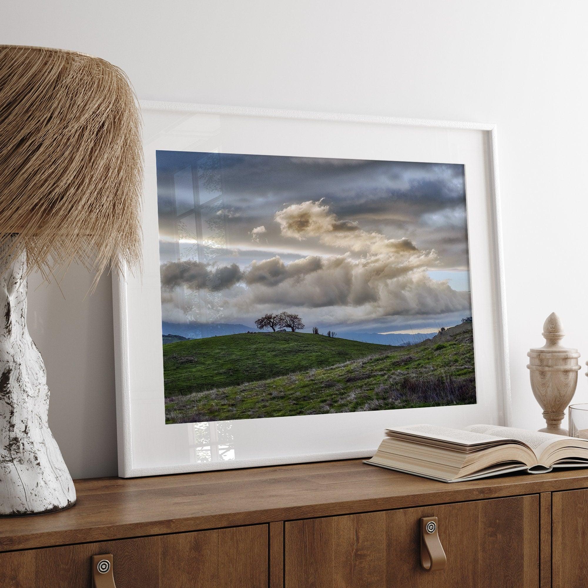 A fine art California moody landscape photography print showing a lone tree standing atop a hill against a dramatic cloudy sky in this scenic landscape wall art.