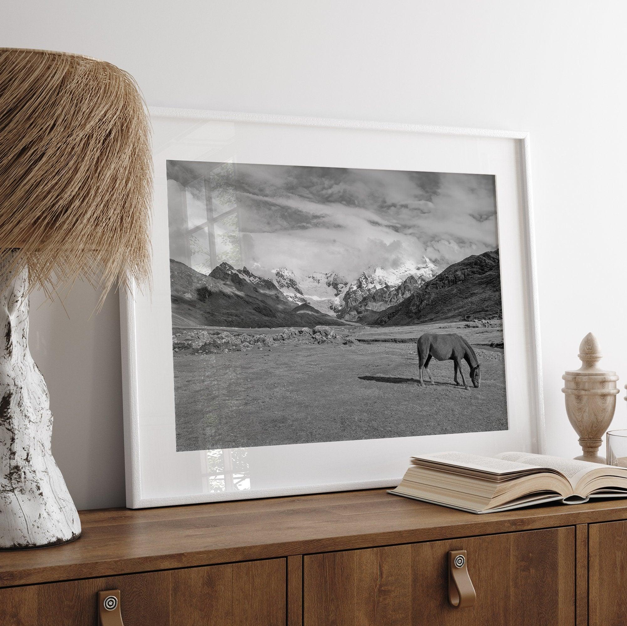 A black and white mountain horse fine art print that can come framed on unframed and was taken in Peru. This mountain wall art showcases snow-covered mountains in Peru and a charming horse grazing relaxingly in the forefront of the picture.