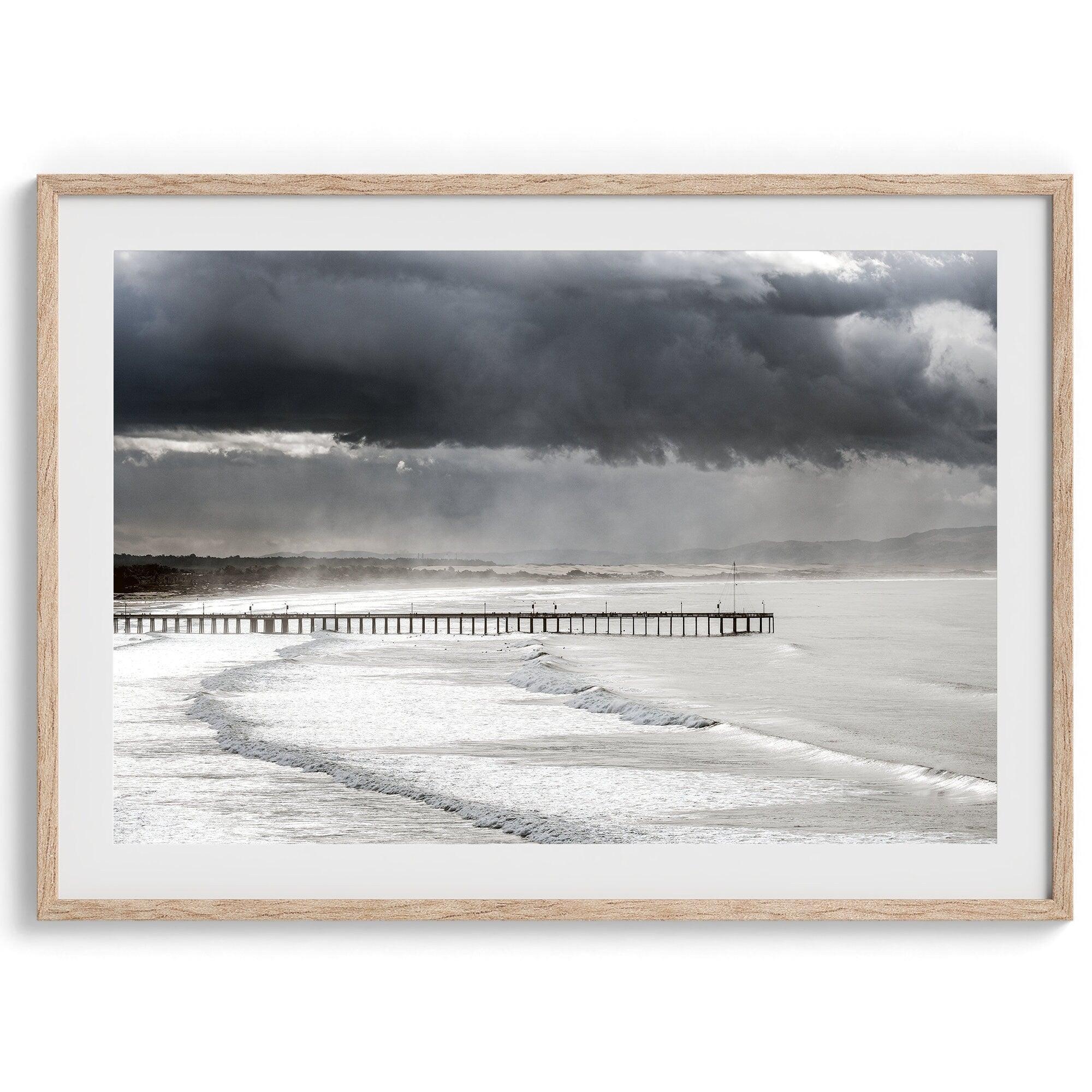 This fine art ocean photography print was taken in Pismo Beach, California, and showcases a distant view of an ocean pier and stormy clouds. Looking long and hard, you can see tens of surfers around the pier.