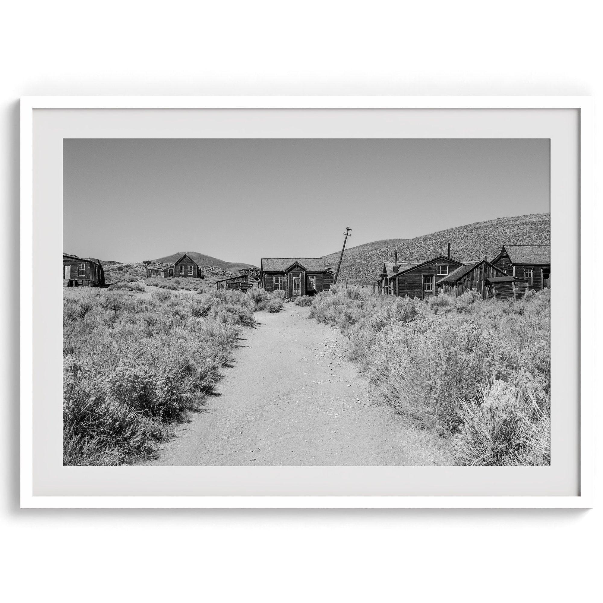 A fine art black and white desert print of a gold rush era town with a road and flowers leading to it. Taken in Bodie State Park, California.