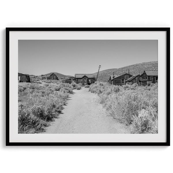 A fine art black and white desert print of a gold rush era town with a road and flowers leading to it. Taken in Bodie State Park, California.