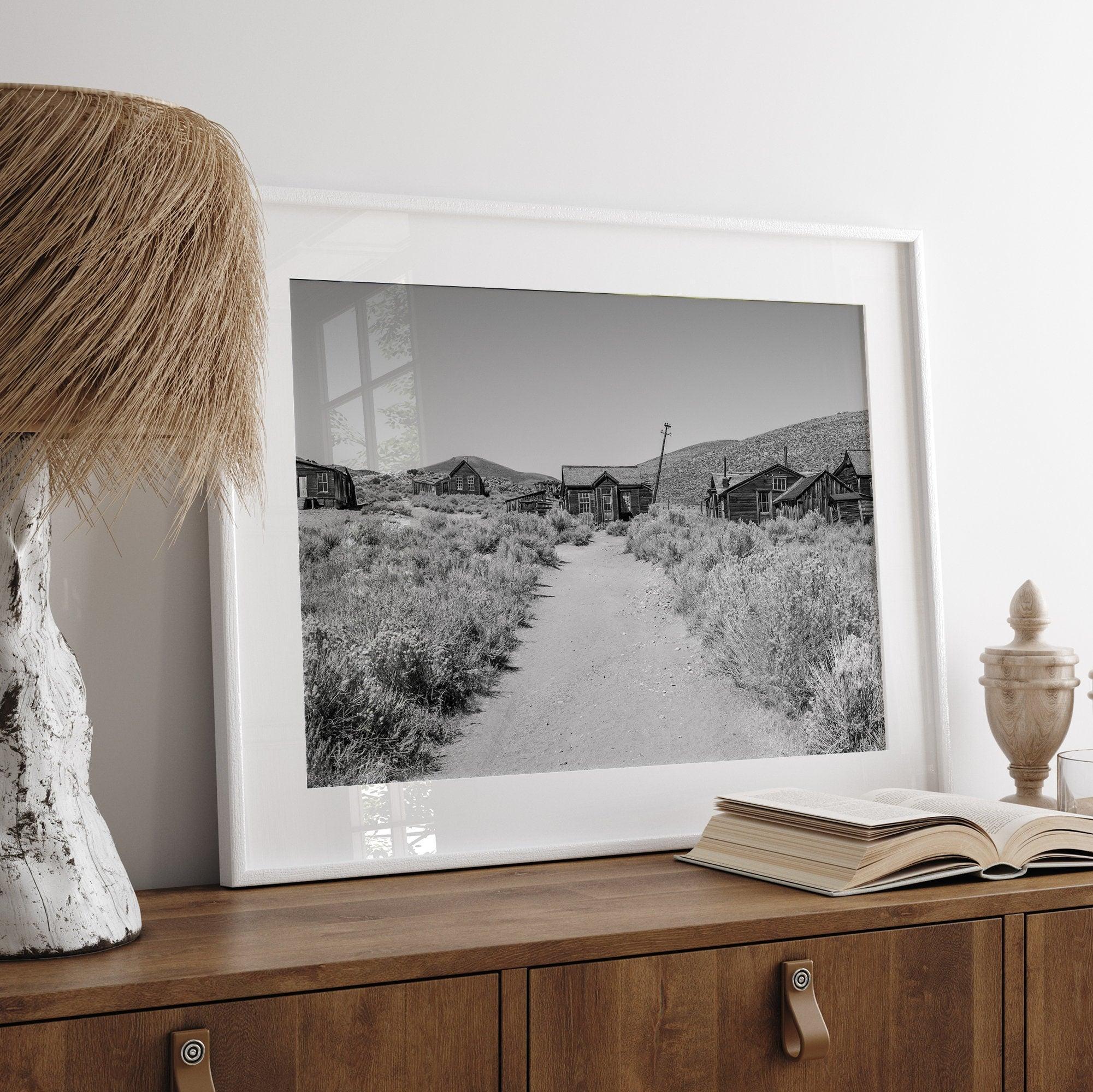 A fine art black and white desert print of a gold rush era town with a road and flowers leading to it. Taken in Bodie State Park, California.