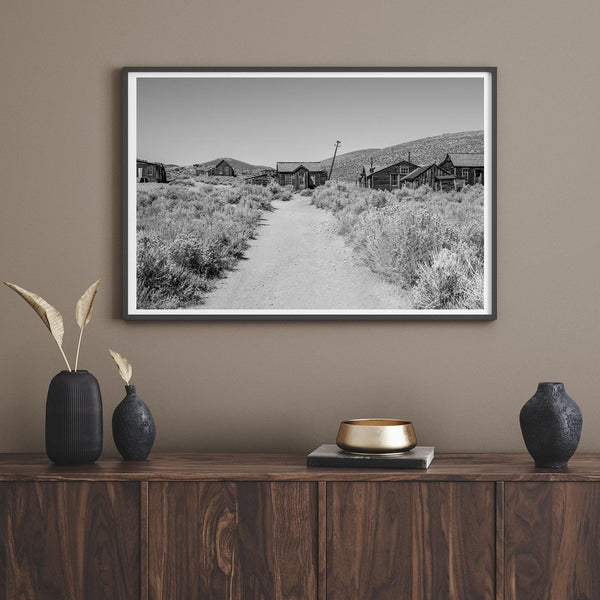 A fine art black and white desert print of a gold rush era town with a road and flowers leading to it. Taken in Bodie State Park, California.