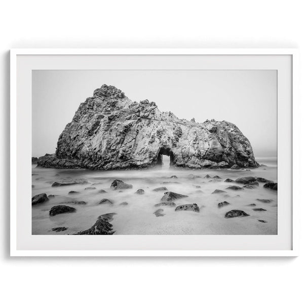 A fine art black and white print of Big Sur Pfeiffer Beach. This beach wall art shows a huge rock on the beach with an opening towards the open sea. This fine art photograph is shot in long exposure so the waves look creamy, gliding over the rocks