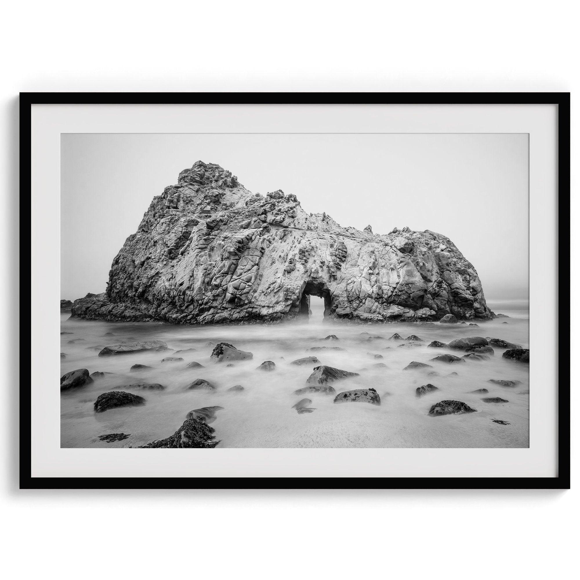 A fine art black and white print of Big Sur Pfeiffer Beach. This beach wall art shows a huge rock on the beach with an opening towards the open sea. This fine art photograph is shot in long exposure so the waves look creamy, gliding over the rocks