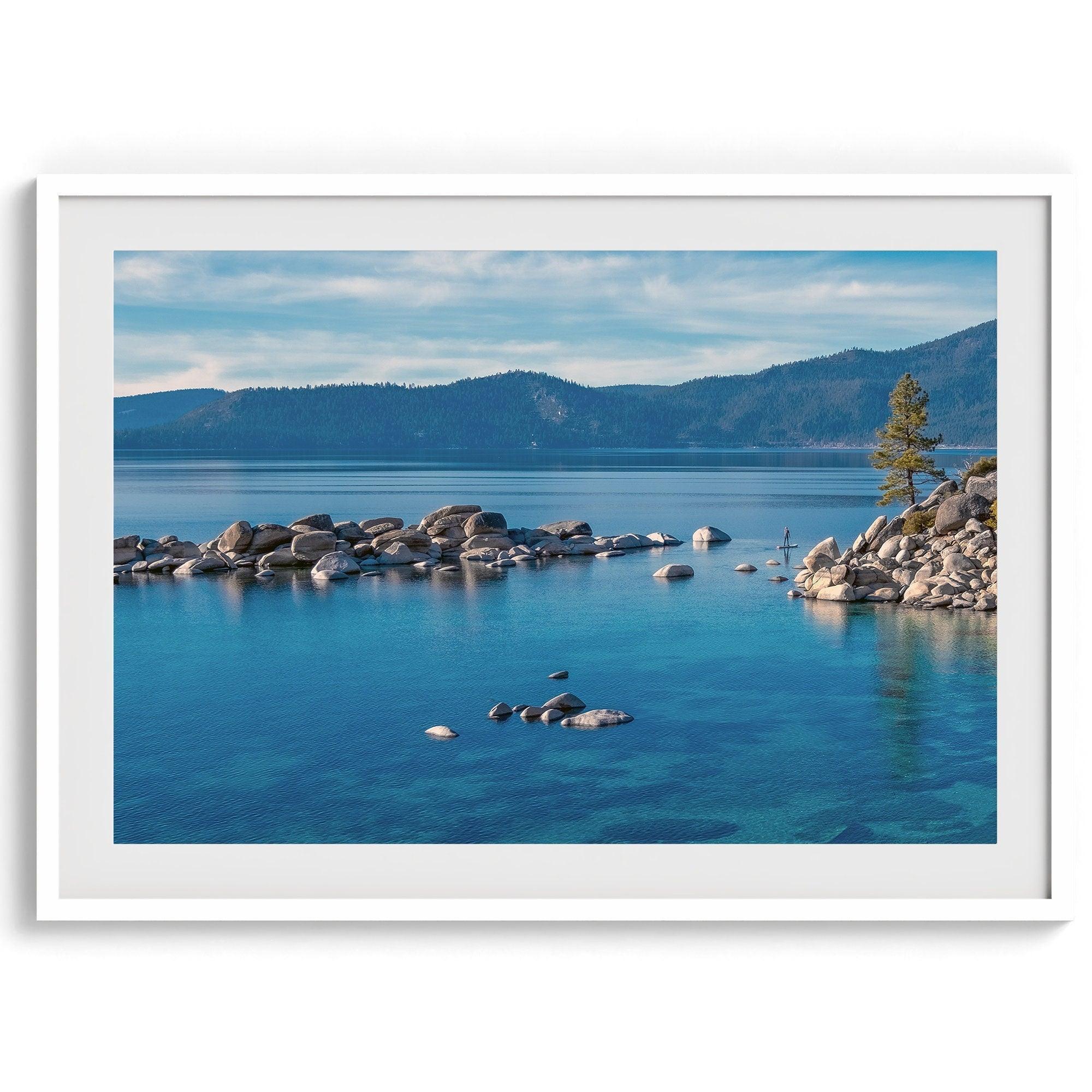 A fine art Lake Tahoe print showcasing a lone stand-up paddle peacefully gliding across the clear water lake near the huge rocks of Sand Harbor Beach with the backdrop of the Sierra Nevada mountains.