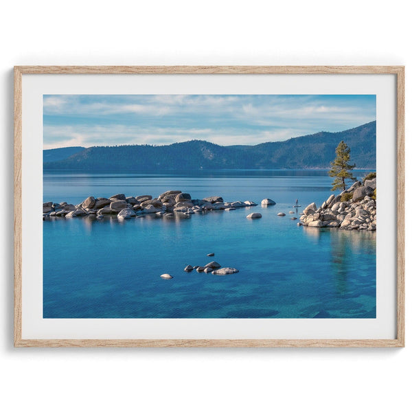 A fine art Lake Tahoe print showcasing a lone stand-up paddle peacefully gliding across the clear water lake near the huge rocks of Sand Harbor Beach with the backdrop of the Sierra Nevada mountains.