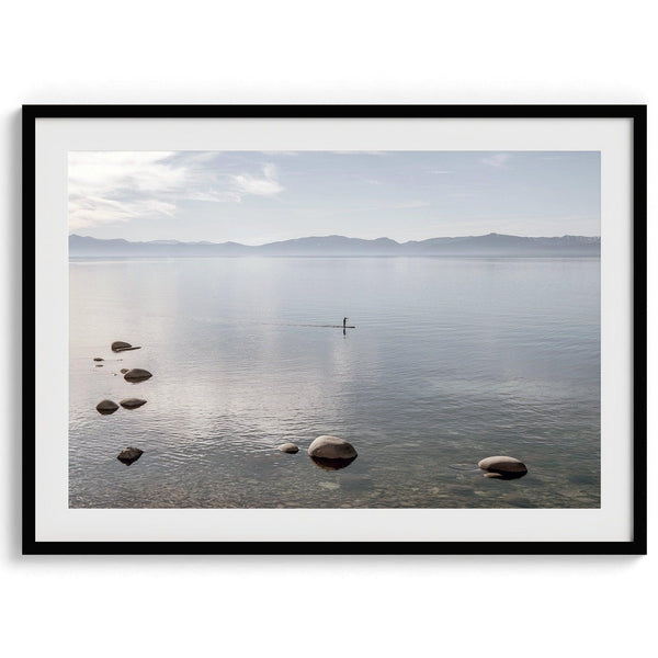Fine art photography print of Lake Tahoe featuring a serene blue water surrounded by Pacific Northwest mountains, with a lone stand-up paddle silhouette on the lake.