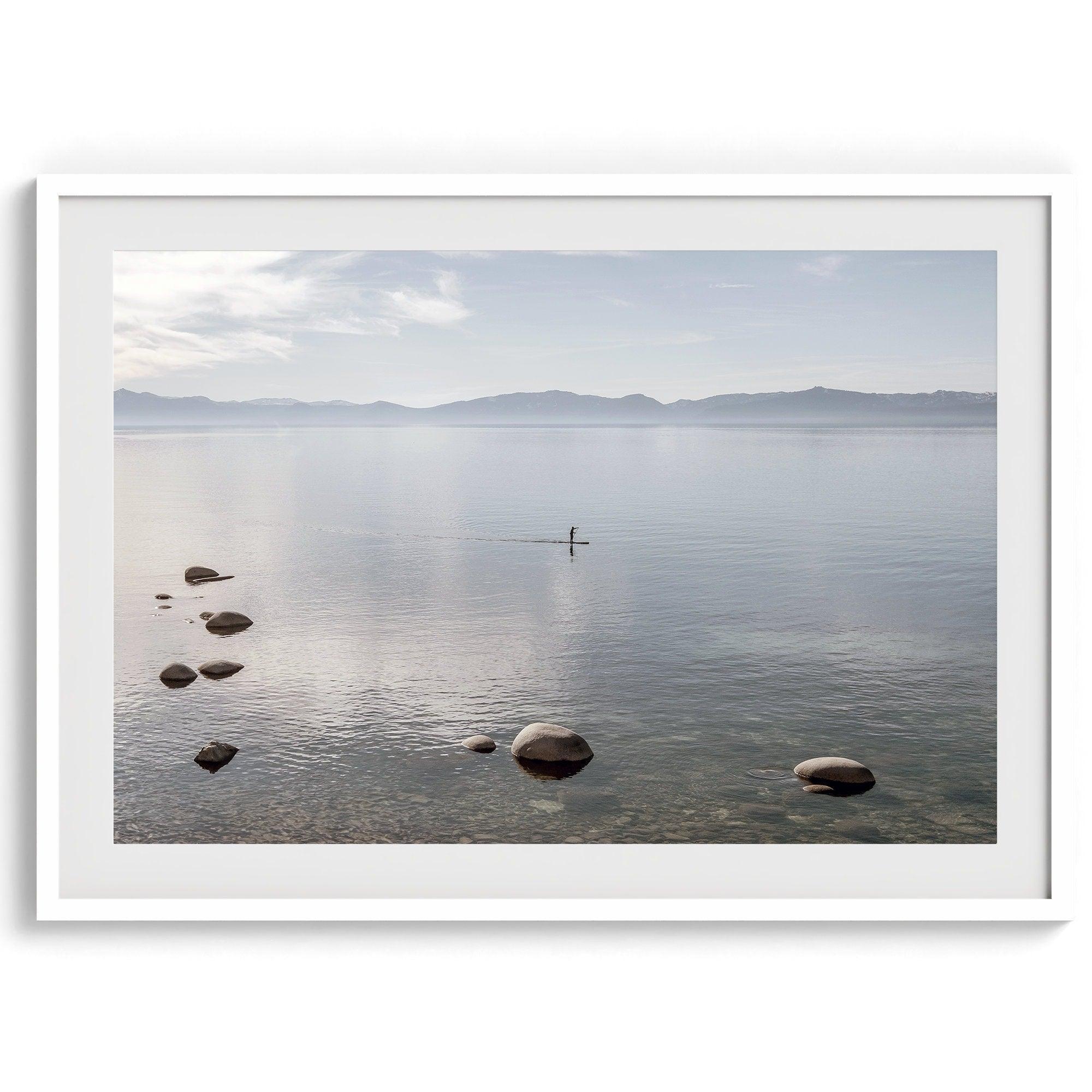 Fine art photography print of Lake Tahoe featuring a serene blue water surrounded by Pacific Northwest mountains, with a lone stand-up paddle silhouette on the lake.