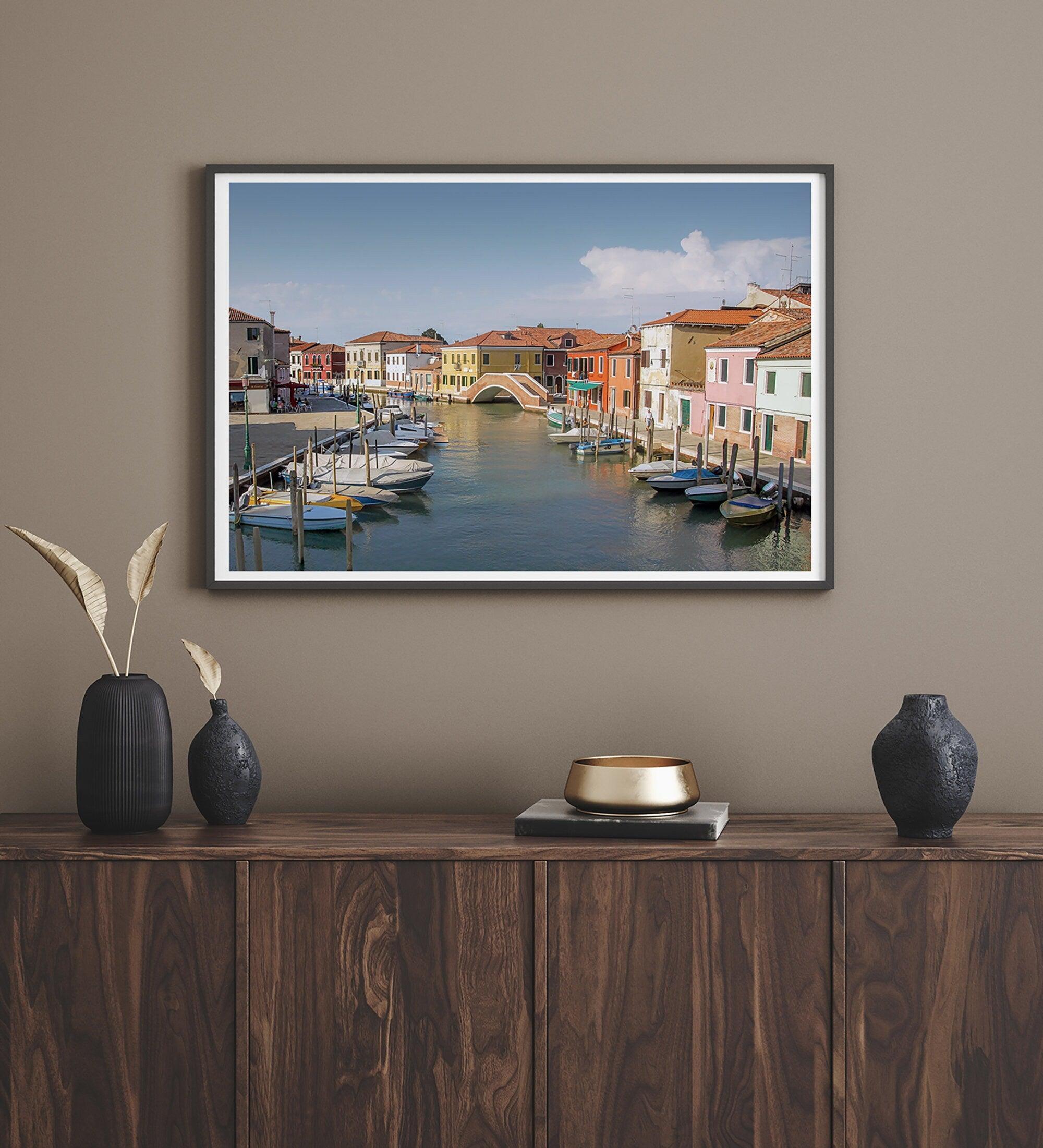 A stunning framed Venice print. The picturesque vibrant streets of Murano are lined with houses in an array of vibrant colors. The sun casts a beautiful glow on the tranquil canal, which adds to the charm of this peaceful Italian town.