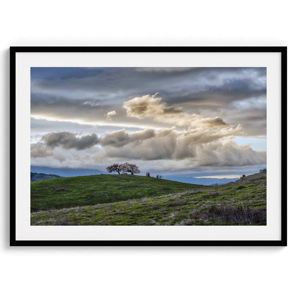 A fine art California moody landscape photography print showing a lone tree standing atop a hill against a dramatic cloudy sky in this scenic landscape wall art.